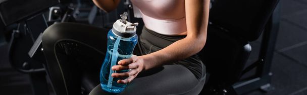 vista recortada de la deportista sentada en la máquina de entrenamiento en el gimnasio y sosteniendo la botella de deportes, pancarta - Foto, imagen
