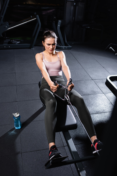 high angle view of athletic sportswoman exercising on rowing machine near sports bottle on floor - Photo, Image