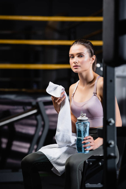 young sportswoman holding sports bottle and towel while sitting on training machine on blurred background - Foto, Imagen
