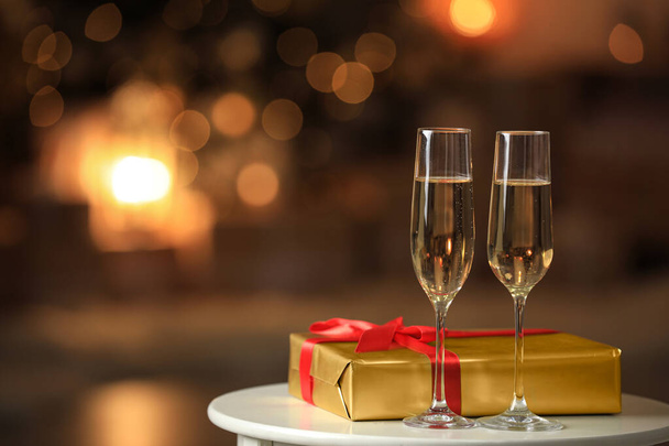 Glasses with champagne and Christmas gift on table in room, bokeh effect. Space for text - Фото, зображення