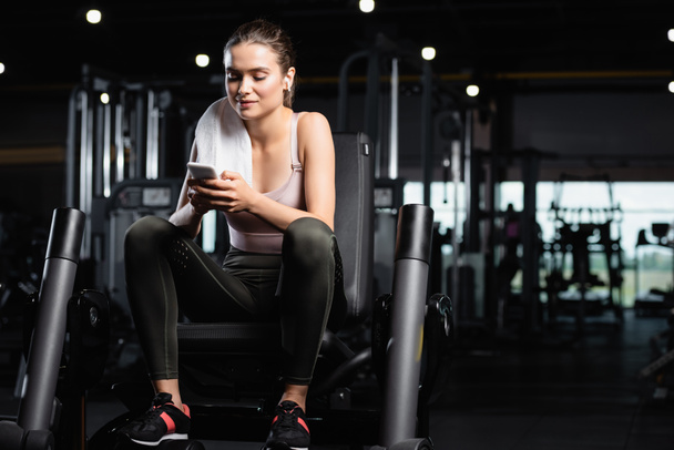 sportswoman sitting on training machine, chatting on smartphone and listening music in wireless earphone - Photo, Image