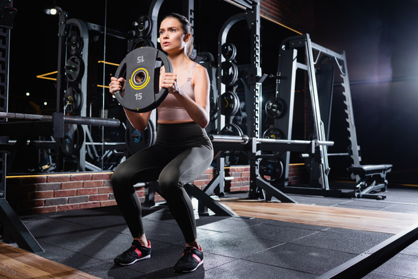 young sportswoman in leggings and sneakers doing sit ups with weight disk in gym - Photo, Image