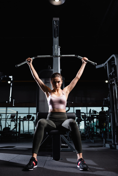 young sportswoman looking at camera while training on lat machine in gym - Photo, Image