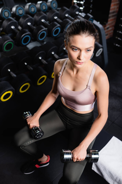 high angle view of sportswoman sitting in gym with dumbbells and looking away - Photo, Image