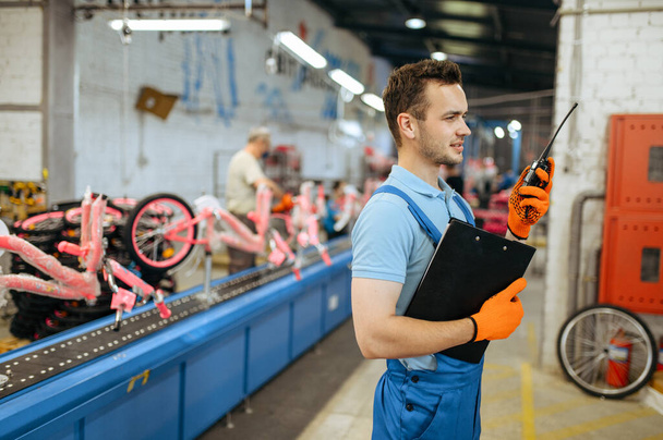 Fábrica de bicicletas, trabajador con un walkie-talkie posa en la línea de montaje de bicicletas. Mecánico masculino en uniforme instala piezas de ciclo en taller, fabricación industrial - Foto, imagen
