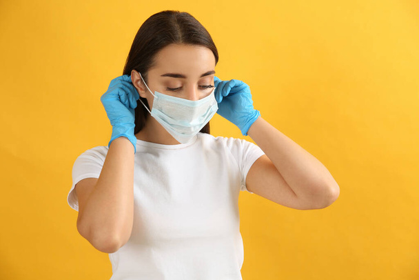 Woman in medical gloves putting on protective face mask against yellow background - Фото, изображение