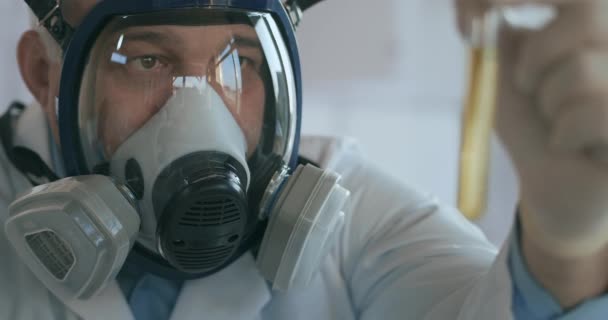 Close up no laboratório um homem em uma tela respiradora de vidro segura um tubo de vidro com um líquido amarelo contra o vírus e olha para ele - Filmagem, Vídeo
