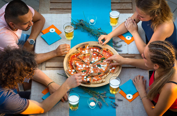 Four young boys sitting at the table on the terrace taking slices of pizza from the plate - Photo, Image