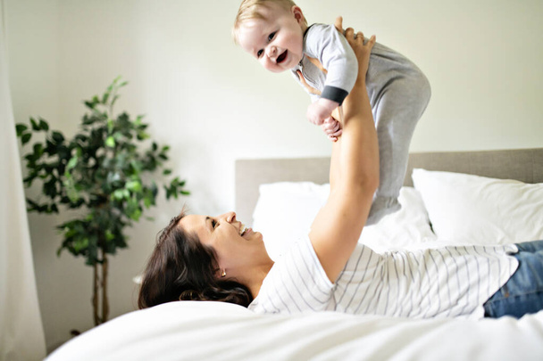 Mother and child on a white bed playing in sunny bedroom - 写真・画像