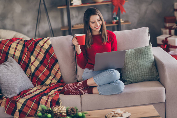 Photo of young charming girl sit sofa hold computer cup toothy smile wear red pullover jeans socks in decorated x-mas living room home indoors - Photo, Image