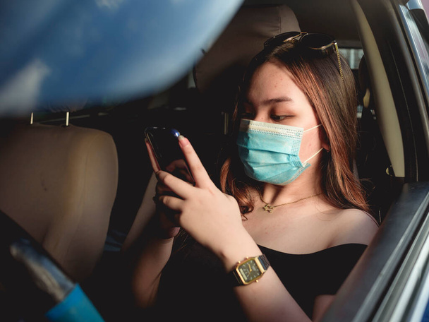 Een jong Aziatisch meisje met een chirurgisch masker is bezig met chatten op sociale media terwijl in de auto geparkeerd bij de garage, parkeerplaats of aan de kant van de weg. - Foto, afbeelding