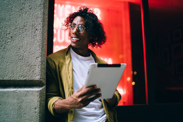 Smiling curly hipster guy in eyewear standing on neon light street with digital tablet share contetnt in vlog,handsome man influencer using touchpad for blogging and share media and multimedia content - Photo, Image
