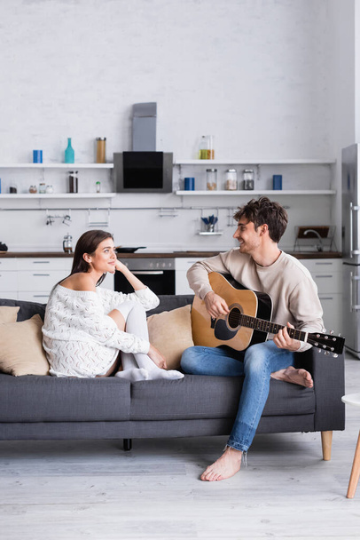 Cheerful couple with acoustic guitar looking at each other on couch  - Foto, immagini