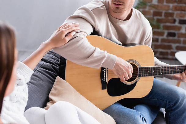 Homem tocando guitarra acústica perto da namorada em primeiro plano desfocado em casa  - Foto, Imagem