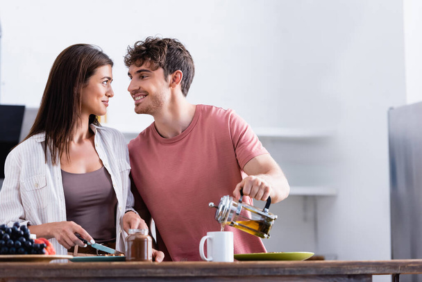 Hombre sonriente mirando a su novia mientras vierte té cerca de frasco de chocolate extendido en primer plano borroso  - Foto, imagen
