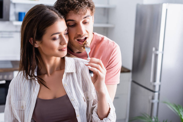 Young woman feeding boyfriend with piece of waffle in kitchen  - Photo, Image