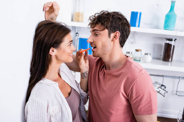Smiling woman feeding boyfriend with slice of paprika near wall in kitchen  - Zdjęcie, obraz