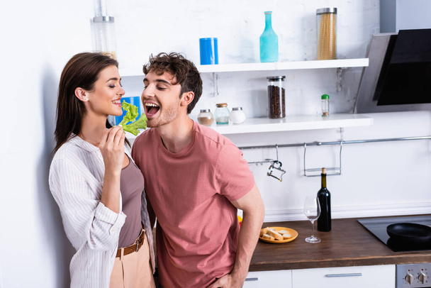 Mujer alegre sosteniendo lechuga cerca de novio con boca abierta en la cocina  - Foto, imagen