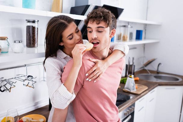 Young woman hugging and feeding boyfriend with baguette in kitchen  - Foto, Bild