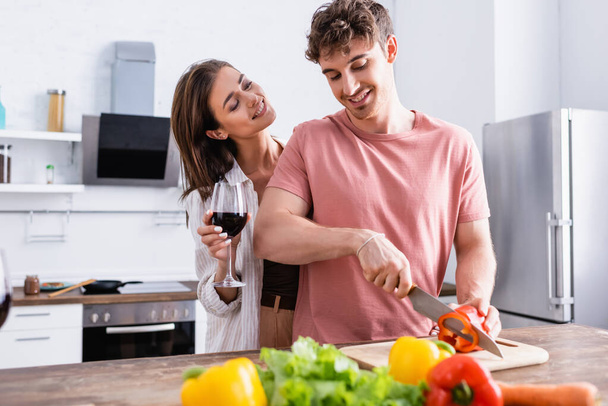 Smiling woman holding glass of wine near boyfriend cutting paprika in kitchen  - Photo, Image
