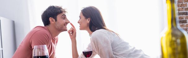 Smiling woman touching nose of boyfriend near wine on blurred foreground, banner - 写真・画像
