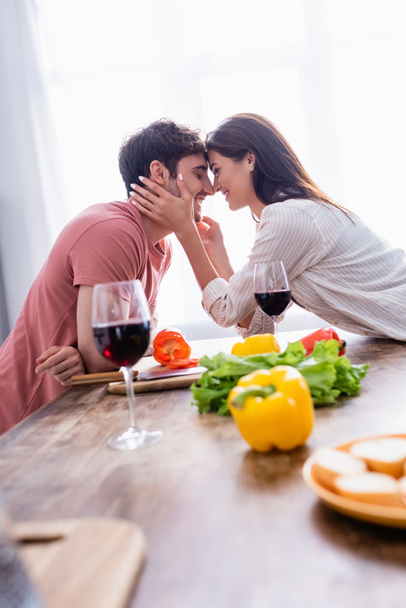 Smiling woman touching face of boyfriend near wine and vegetables on blurred foreground  - 写真・画像