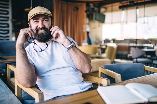Positive bearded male with glasses wearing in casual clothes smiling and speaking with coworker on mobile phone during work in modern creative cafe - Photo, Image