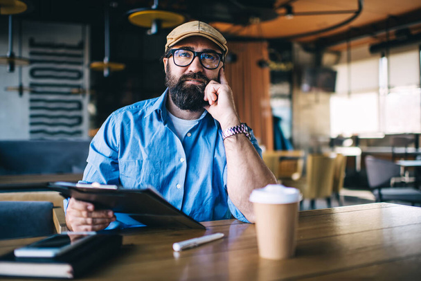 Thoughtful mature bearded male wearing eyeglasses blue shirt and flat cap sitting in cafe with notepad leaning on hand and contemplating about problem while looking away - Fotografie, Obrázek