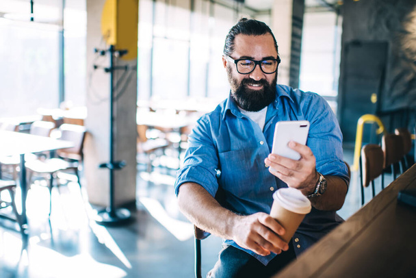 Happy confident mature male entrepreneur in casual wear sitting near bar station in cafe, cheerful hospitable bearded man owner of small business coffee shop checking notification on smartphone - Фото, зображення