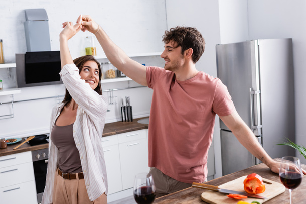 Smiling man dancing with girlfriend near paprika and wine on blurred foreground in kitchen  - Fotó, kép