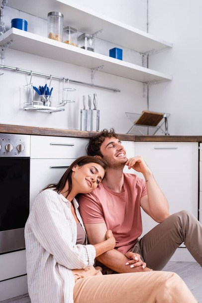 Smiling woman with closed eyes sitting near boyfriend on floor in kitchen  - Fotografie, Obrázek