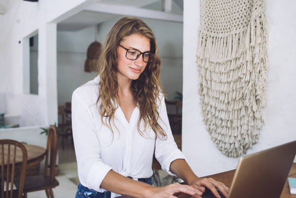 Smiling thoughtful adult female in glasses and casual clothes focusing on screen and interacting with laptop while standing at counter in cafe - Foto, Bild