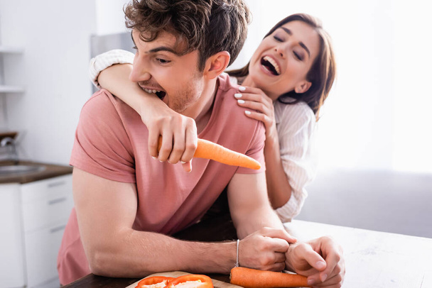 Man biting hand of cheerful girlfriend holding carrot in kitchen  - Foto, imagen