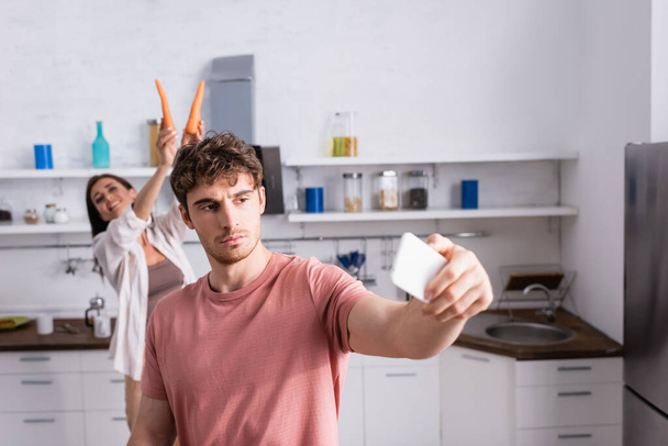 Man taking selfie with smartphone near cheerful girlfriend with carrots on blurred background  - Foto, immagini