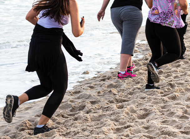 Vista trasera mirando hacia abajo a algunas mujeres caminando y corriendo en el borde de la playa cerca del agua usando spandex en la tarde durante el verano 3. - Foto, imagen