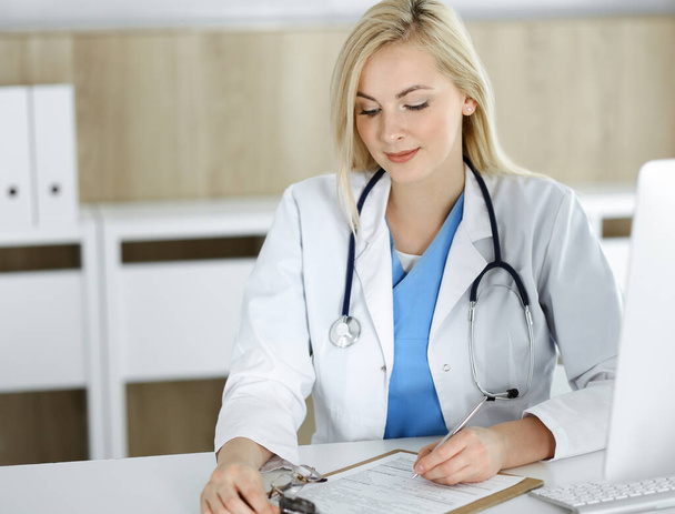 Woman-doctor at work while sitting at the desk in hospital or clinic. Blonde cheerful physician filling up medication history record form - Photo, image