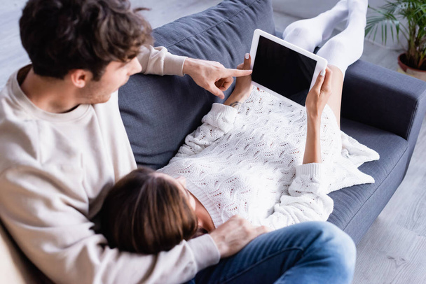 Mężczyzna wskazując na cyfrowy tablet z pustym ekranem obok dziewczyny na kanapie na niewyraźnym pierwszym planie  - Zdjęcie, obraz