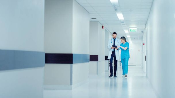 Surgeon and Female Doctor Walk Through Hospital Hallway, They Consult Digital Tablet Computer while Talking about Patients Health. Modern Bright Hospital with Professional Staff. - Foto, Imagem