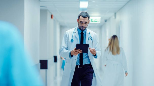 Determined Handsome Doctor Uses Digital Tablet Computer while Walking Through Hospital Hallway. Modern Bright Clinic with Professional Staff. - Foto, Imagen