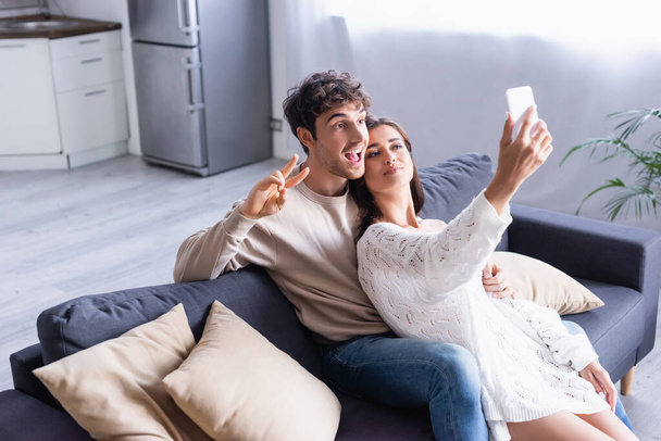 Cheerful man showing peace sign near girlfriend taking selfie on smartphone on couch  - Fotografie, Obrázek