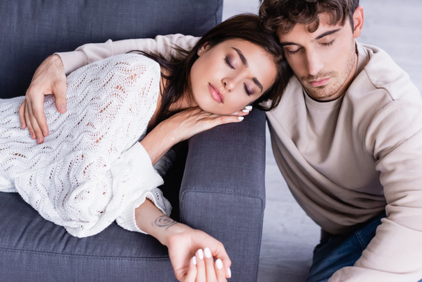 Junger Mann umarmt brünette Freundin mit geschlossenen Augen auf Couch  - Foto, Bild