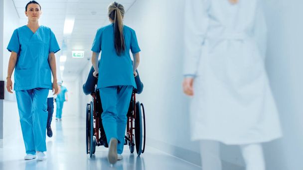 Shot of the Female Nurse Moving Patient in the Wheelchair Through the Hospital Corridor. Doing Procedures. Bright Modern Hospital with Friendly Staff. - Фото, изображение
