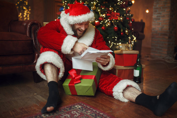 Slechte dronken Kerstman leest brieven onder kerstboom, smerig feest, humor. Ongezonde levensstijl, man met baard in vakantiepak, nieuwjaar en alcoholisme - Foto, afbeelding