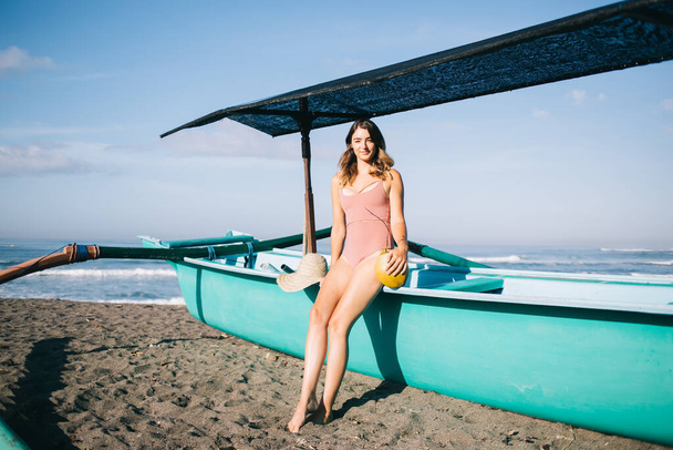 Portrait of female posing near water boat at tropical sand beach with coconut fruit and straw hat enjoying sunbathing leisure, attractive Caucasian woman looking at camera during solo vacations - Foto, Imagem