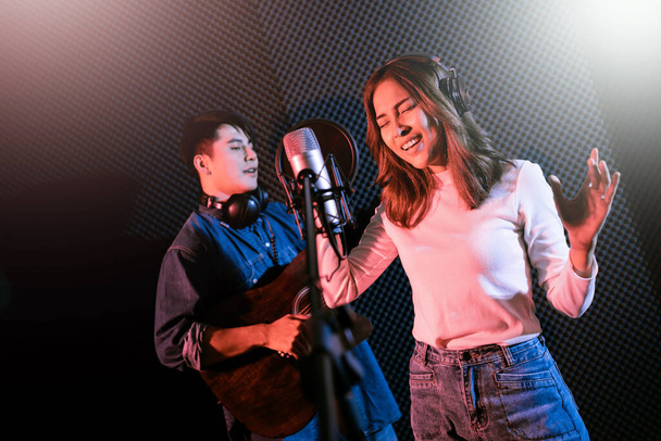 Pretty Asian female singer recording songs by using a studio microphone and pop shield on mic with male playing guitar in blue and red light. Performance and show in the music business. Duet session. - Foto, Imagem