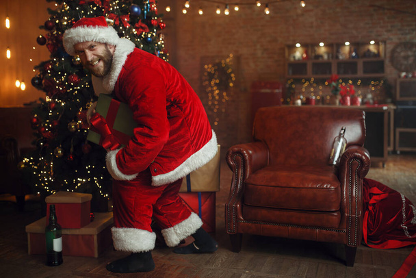 Zlý opilý Santa Claus krade dárky pod vánočním stromečkem, ošklivou párty, humor. Nezdravý životní styl, vousatý muž v svátečním kostýmu, nový rok a alkoholismus - Fotografie, Obrázek