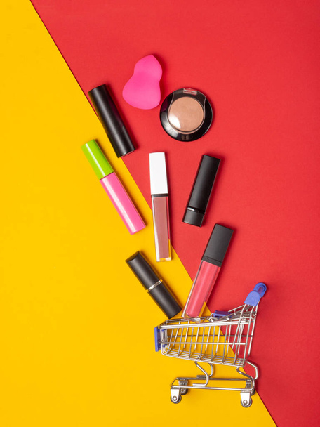 A set of lipstick cosmetics and lip glosses, powder, eyeshadow and a shopping trolley on a bright red and yellow background. The concept of buying cosmetics, online shopping, holiday - 写真・画像