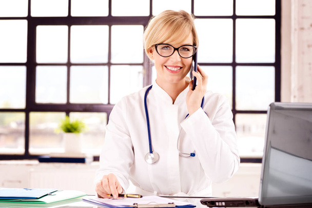 Portrait shot of smiling female doctor sitting at desk and making a call with her patient while working in doctors office. - Foto, Imagen