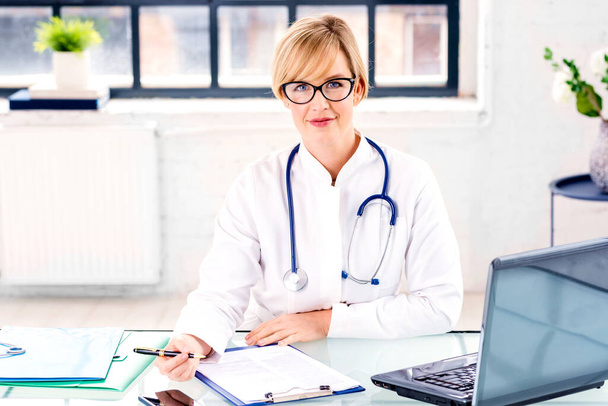 Retrato de una doctora sonriente sentada en el escritorio y trabajando en el consultorio médico. - Foto, imagen