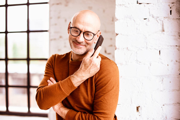 Portrait shot of happy senior man wearing casual clothes while standing by the window and talking with somebody on his mobile phone.  - Fotoğraf, Görsel